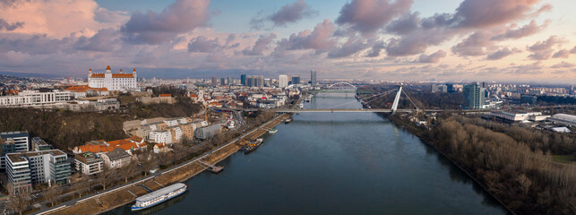 Aerial view of Bratislava, Slovakia, shows the Danube, Old Town, and modern districts at sunset. Bratislava Castle and the UFO Bridge span the scene with boats and bare trees.