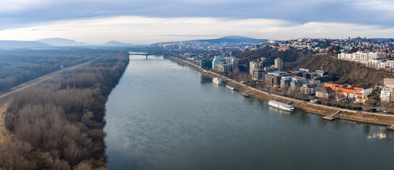 Fototapeta premium Aerial view of Bratislava, Slovakia, along the Danube River, with riverside developments and hillside neighborhoods right, wooded floodplains left, bridge and riverboats visible.