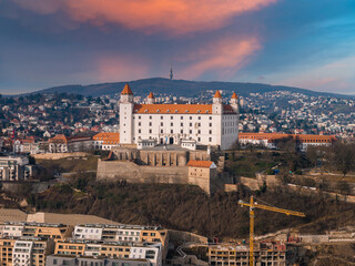 Fototapeta premium Aerial view of Bratislava Castle at dusk, white fortress with four towers, terraces and ramparts, Kamzik TV Tower on ridge, cranes and new buildings in foreground.