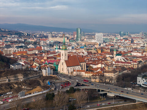 Aerial view of Bratislava, Slovakia, with St. Martin's Cathedral, terracotta rooftops, a multi level highway interchange, and modern high rises under cool winter haze.