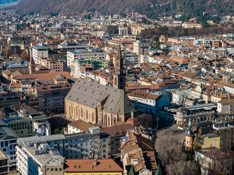 Aerial view of Bolzano, Italy, shows Bolzano Cathedral with patterned roof and spire, pastel and terracotta roofs, wooded hills, and crisp midday winter light.