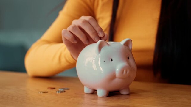 A woman with long dark hair is adding coins into a white piggy bank on a wooden table. The setting features a simple and modern background, focusing on the activity of saving money.