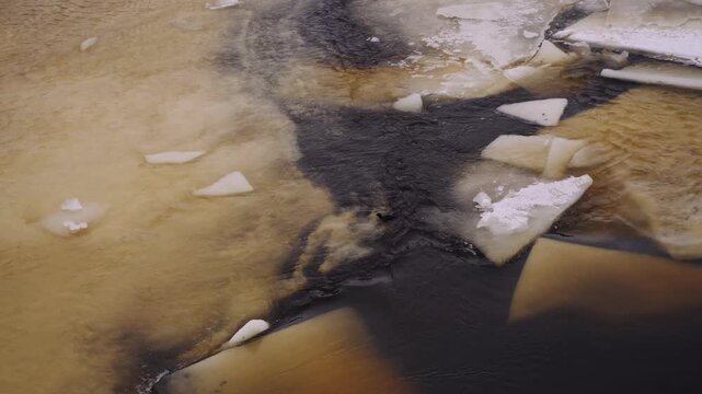 Aerial view of river currents merging through an ice bottleneck in cinematic slow motion. A high-resolution 4K study of dark, tannin-rich water weaving between large stationary ice sheets