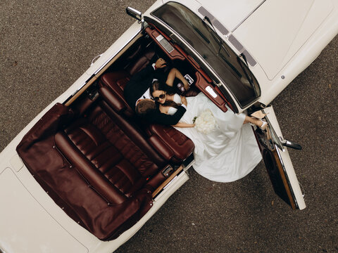 Aerial top down view of a fashionable wedding couple relaxing in a vintage convertible car with red leather interior. Bride in elegant white dress and groom in sunglasses enjoying romantic road trip. 