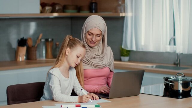 A woman wearing a headscarf helps her daughter with schoolwork at a kitchen table. The daughter draws with markers while the mother uses a laptop to assist. Bright daylight comes through the window.