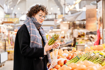 Curly haired white woman choosing asparagus at indoor market produce stall