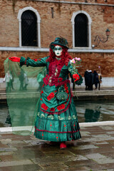 Naklejka premium Venice, Italy - February 09, 2026: Woman in elaborate green costume with mask and flowers poses during Venice Carnival near canal and historic brick buildings
