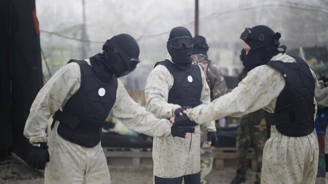 A group of paintball players in protective gear performs a team hand stack and fist bump before a game. Individuals in camouflage and masks demonstrate unity and readiness for competition.