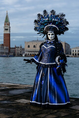 Obraz premium Venice, Italy - February 09, 2026: Woman in elaborate blue costume and mask poses at Venice Carnival with historic buildings and bell tower in the background