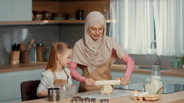 A woman with light skin and a girl are enjoying a baking session in a modern kitchen. The woman is teaching the girl to roll dough. They are smiling and engaged in the activity.