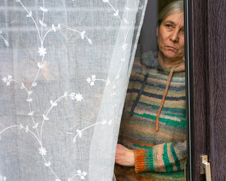 Serious mature woman looking out of window from behind curtain at home