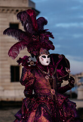Obraz premium Venice, Italy - February 09, 2026: Elegant masked figure in ornate purple costume with feathers at Venice Carnival, historic architecture in the background