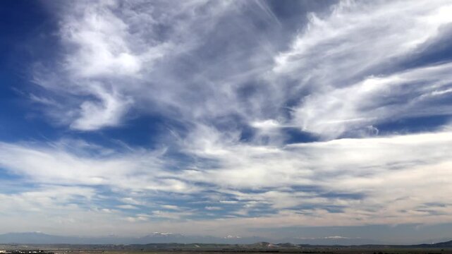 Timelapse storm clouds and cirrus drift across blue sky, revealing fast changing weather. Accelerated cloudbank flow shows swirling vapor layers, shifting light, unstable atmosphere, moody heavens.