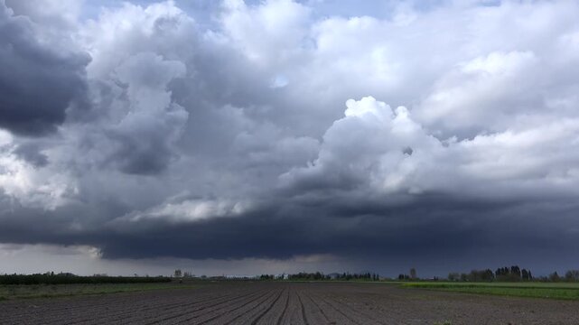 Storm clouds timelapse above plowed field as tractor works on broad countryside plain. Dramatic thunderhead motion clears after rain, furrowed farmland and machinery under shifting sky.