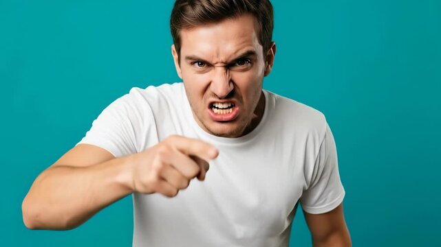 Intense angry young man pointing aggressively against teal backdrop