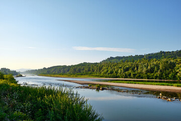 Obraz premium Peaceful Prut river flowing through a green valley at sunrise. Warm sunlight illuminates forested hills and calm water, creating a scenic summer landscape with tranquil natural beauty