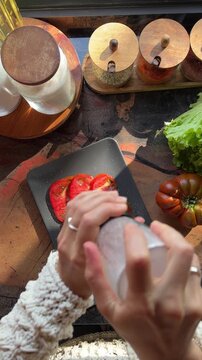 Top view POV of a woman seasoning fresh caprese salad in bright natural sunlight. Healthy Italian food preparation with tomatoes, mozzarella. Clean home cooking lifestyle, summer meal concept