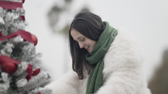 A teenage girl joyfully decorates a festive Christmas tree adorned with red ribbons and ornaments. She smiles while adding finishing touches to the winter holiday decoration outdoors.