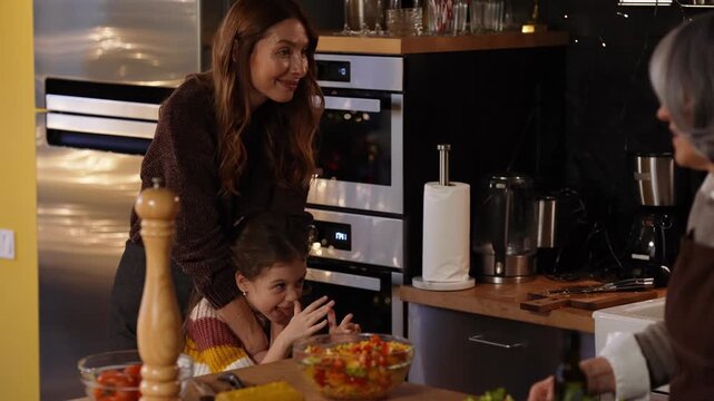 A mother hugs her daughter who is making faces at her brother while their grandmother stands next to them and they talk