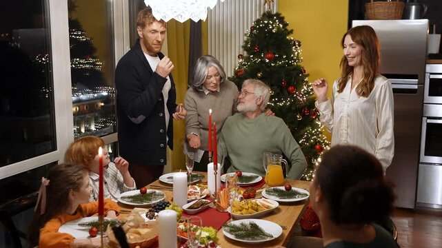 The father lights a candle with a match while the family and guests sit at the table and the mother and grandmother stand next to him and they talk and laugh