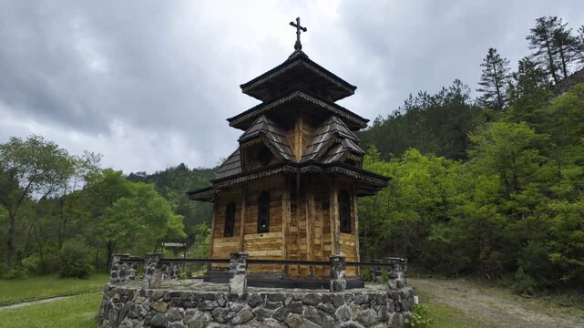 Traditional wooden church Jatarice in Mokra Gora surrounded by green forest and mountains under cloudy sky. Historic architecture landmark in peaceful rural Serbia landscape