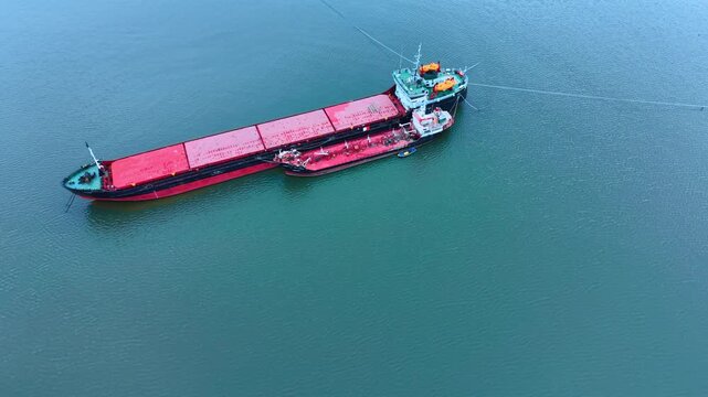 Aerial view of a cargo ship and tanker in open water. A large red and black cargo vessel positioned next to a smaller red tanker ship on calm green water.