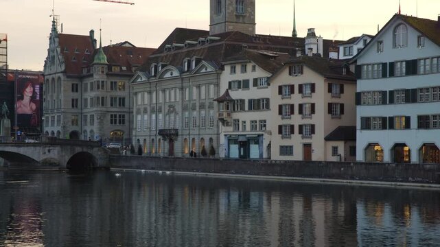 Static shot of historic guild houses and traditional buildings along the Limmat river in Zurich, Switzerland, with their facades reflected in the calm water at dusk.