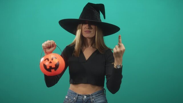 Woman in witch hat holds pumpkin bucket and shows middle finger, crop top midriff visible in studio; playful defiance.