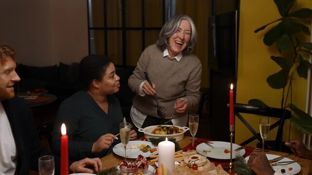A grandmother stands and places food on a plate, in front of which a female guest sits at the table, and they laugh while talking