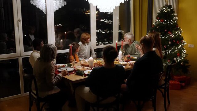 A grandfather sits at a table with his family and guests and listens to his grandson who stands next to them