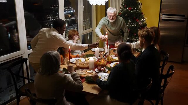Family and guests sit at the table and clink glasses while the grandfather and the male guest stand up