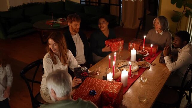 Family and guests sit at a table and talk while granddaughter gives a gift to grandfather and grandson gives a gift to grandmother