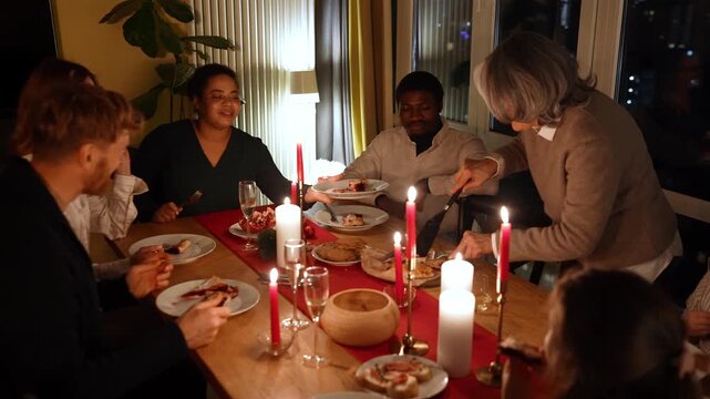 Family and guests sit at the table and hold plates into which the grandmother places pieces of pie, which she cuts while standing