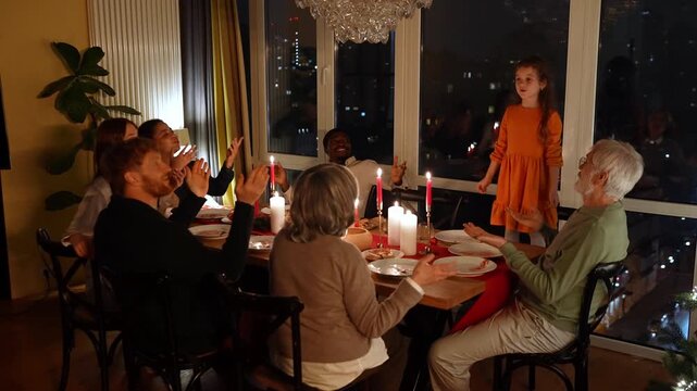 Granddaughter stands on a chair and sings while her family and guests sitting next to her at the table applaud her