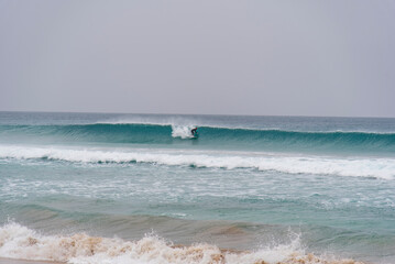 Surfer riding a turquoise Atlantic wave at Playa de la Pared, Fuerteventura, Canary Islands, with white foam and rolling swell under a cloudy sky.