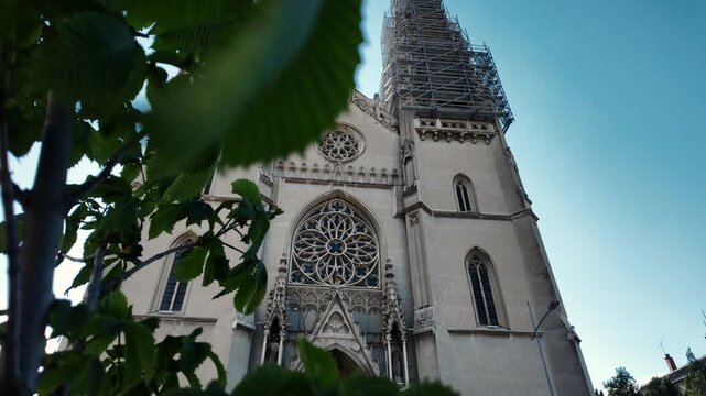 Low angle view of the Catholic cathedral in Vrsac, Serbia framed by green leaves. Gothic facade with large rose window and tower under restoration with scaffolding