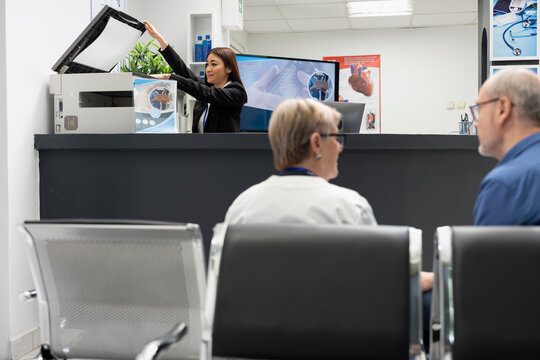 Asian receptionist using copier and scanning machine at front desk of modern healthcare facility. Female hospital secretary handles printing of medical forms as elderly doctor consults with patient.