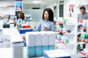 Asian woman stands in pharmacy aisle holding beauty product while comparing details on shelves. Adult female consumer reviews over the counter medication, making careful healthcare purchase decision. © DC Studio