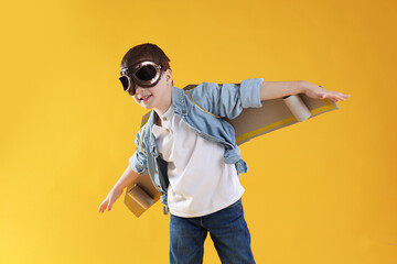 Happy little boy in goggles with cardboard plane wings playing pilot on yellow background