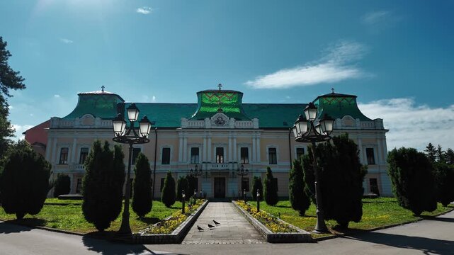Vladicanski dvor with its distinctive green roof and symmetrical facade. Historic bishop palace surrounded by garden, trees and classic street lamps on a sunny day.
