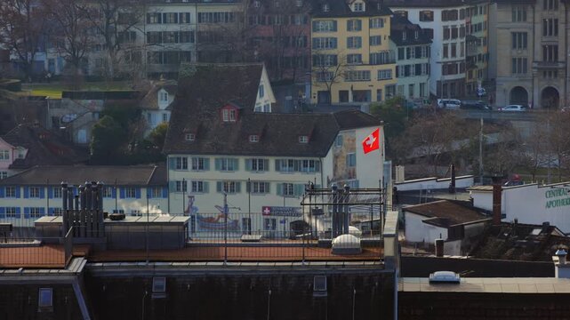 Static shot of the historic old town in Zurich, Switzerland, featuring the iconic "Zunfthaus zur Schiffleuten" with its traditional mural, a waving Swiss flag, and smoking chimneys on a sunny day.