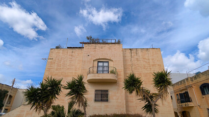 Natural limestone apartment facade with small curved balcony framed by palm trees, bright calm Malta street scene