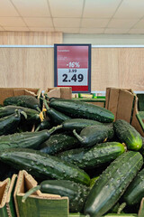 Closeup pile of fresh cucumbers in cardboard crates with price tag and discount percentage in grocery store, consumer inflation mood
