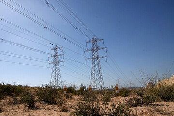High-voltage lattice transmission tower standing in a vast desert landscape, supporting overhead power lines under a clear blue sky. © fornax