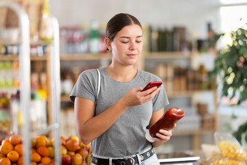 Fototapeta premium Young woman buyer scanning qr code for tomato ketchup sauce in grocery store
