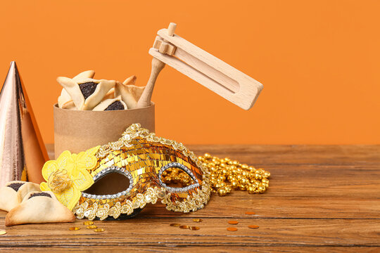 Purim holiday still life with hamantaschen cookies, wooden grogger noisemaker, golden carnival mask, party hat and beads on wooden table against orange background