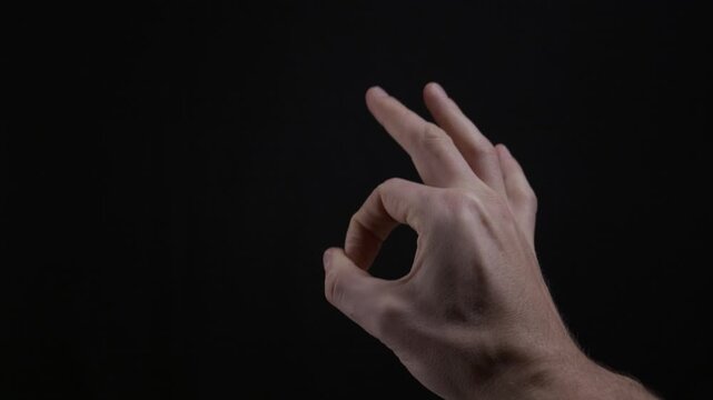 A man's hand makes the A-OK symbol against a solid black background in a studio setting. Concept of approval, perfection, non-verbal communication, satisfaction, and positive agreement.