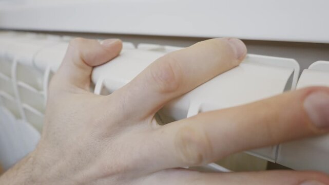 Close up of a male hand touching a white central heating radiator to check for warmth. Concept of the winter heating season, home comfort, energy costs, and utility bills.