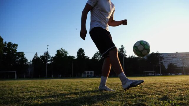 Professional footballer juggling soccer ball on stadium at sunset. Young man kicking ball at green field. Sportsman practicing tricks at meadow with sunlight at background. Freestyle football concept