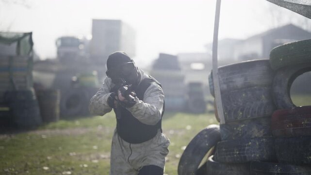 A player in tactical gear aims a paintball rifle in an outdoor arena. He moves strategically and takes a firing position among tire barricades.